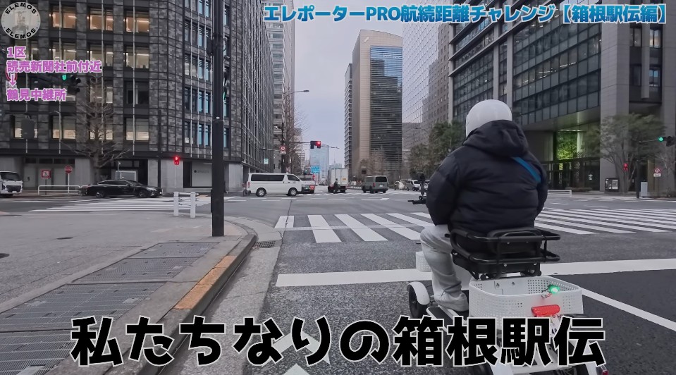 2026-05-04_161116 | ELEMOs Person wearing a white helmet rides a mobility scooter through a city crosswalk, with tall office buildings on both sides and Japanese text overlays top and bottom of the frame.