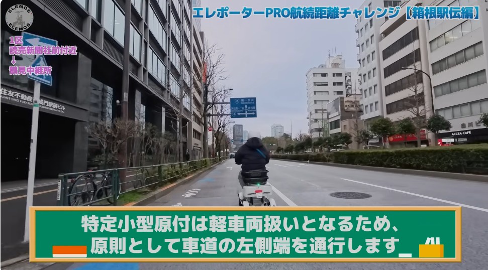 2026-05-04_161611 | ELEMOs City street view with a person riding a scooter in the center lane, tall buildings on both sides; blue video title text at top and a green banner with Japanese instructions across the bottom.
