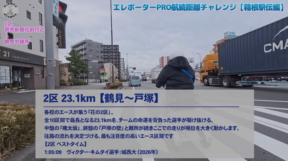 2026-05-04_163650 | ELEMOs City street view with a cyclist in a black jacket riding ahead; a large orange truck drives on the right, and a translucent caption panel covers the lower half with a 23.1km route description in Japanese.
