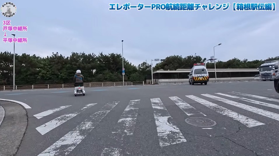 2026-05-04_170906 | ELEMOs Person on a small electric scooter crossing a wide crosswalk at an intersection; an ambulance and a truck on the road ahead, with trees and a blue sky and Japanese banner text at the top.
