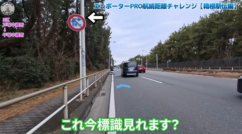 2026-05-04_171120 | ELEMOs Japanese road scene with blue directional arrow in the lane, a circular sign on a pole to the left, cars ahead, and green/blue captions reading 'これ今標識見れます?' (Can you read the sign now?)