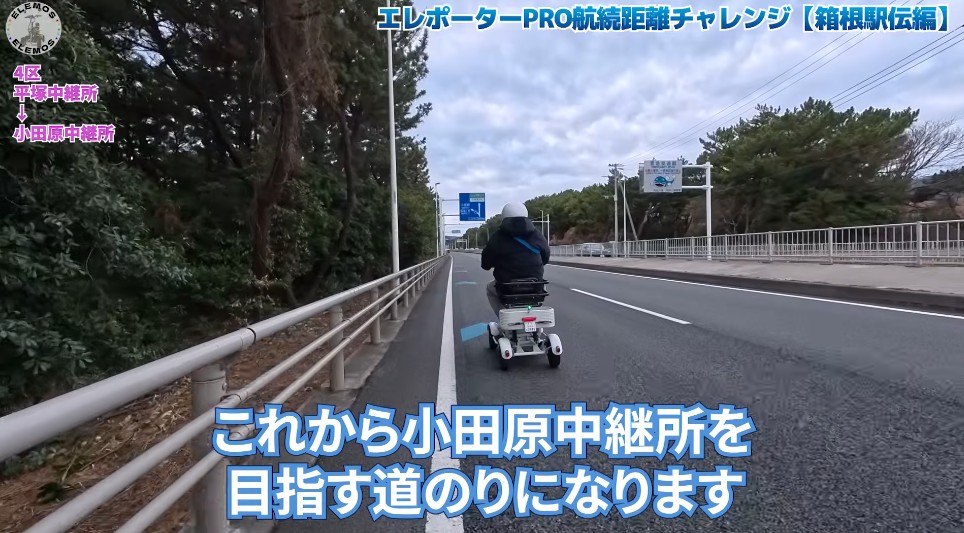 2026-05-04_172019 | ELEMOs Person on a white mobility scooter riding along a road with guardrails and trees, overlaid with blue Japanese text announcing the Odawara relay point journey.