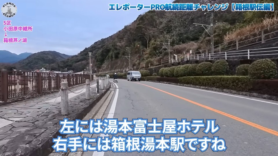 2026-05-04_180431 | ELEMOs Road along a hillside highway with a stone-paned sidewalk on the left and trimmed hedges on the right; a white van and a motorcycle drive toward the viewer, with blue overlay text naming nearby hotel on the left and Hakone Yumoto Station on the right.