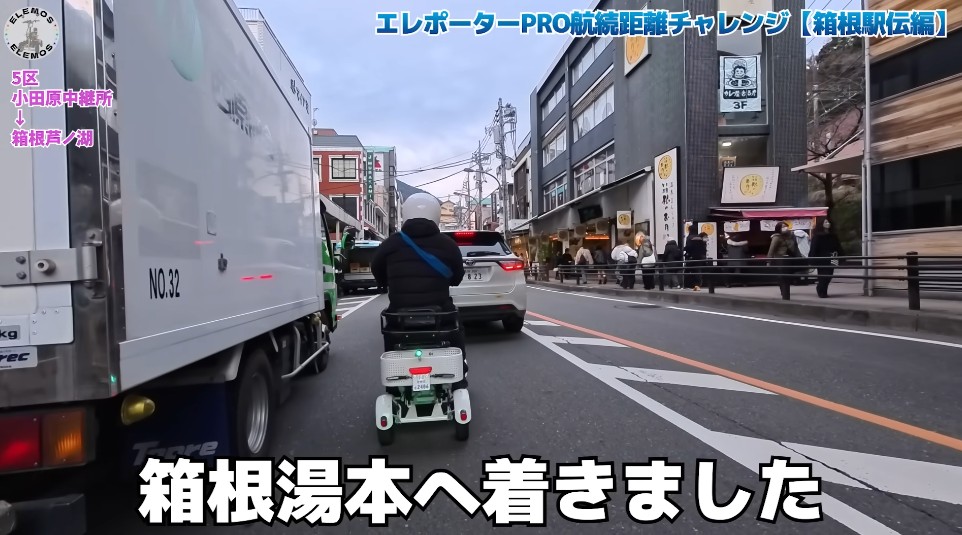2026-05-04_180610 | ELEMOs Person on a small white mobility scooter follows a large truck down a busy street lined with shops; caption says 'We arrived at Hakone-Yumoto' (Hakone-Yumoto Station area).
