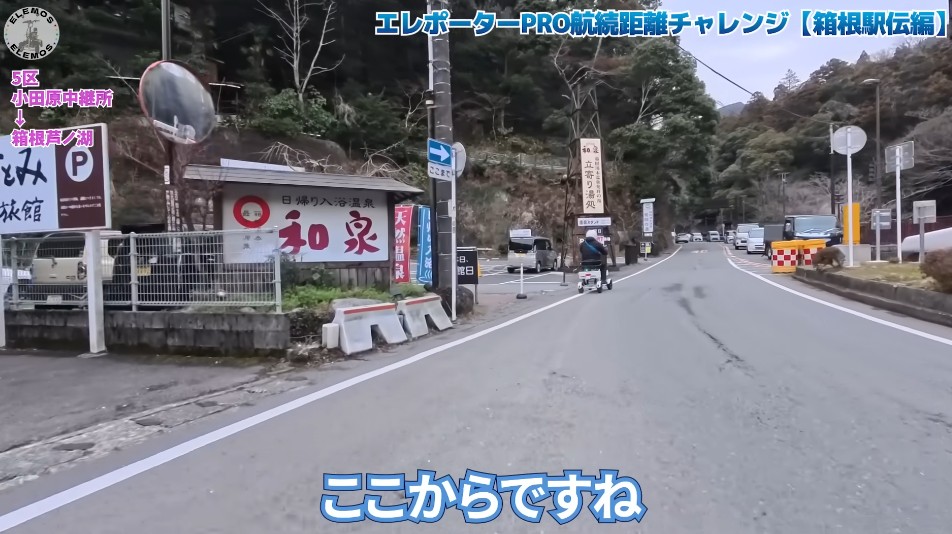 2026-05-04_181027 | ELEMOs Rural Japanese street with a scooter rider approaching, shop signs on the left and forested hills in the background; blue caption overlays on screen.