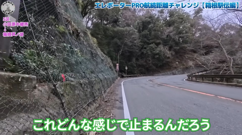 2026-05-04_182010 | ELEMOs Curving mountain road bordered by a metal fence on the left and a guardrail on the right, with trees and rocky embankment; video overlay text in Japanese appears on screen.