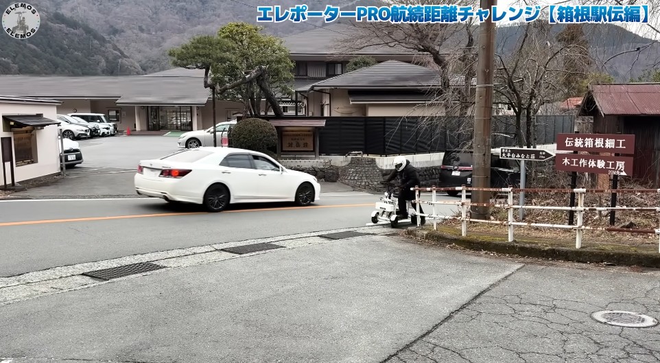 2026-05-04_182313 | ELEMOs White car drives along a rural Japanese street while a person on a small scooter stands by a fence near traditional buildings and distant mountains.