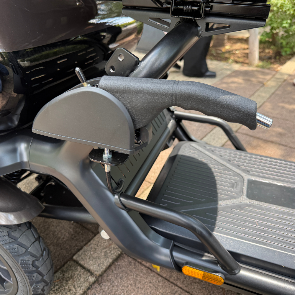Close-up of a scooter's left handlebar and footboard, showing the grip, brake lever, and a gray housing with a bolt and wiring.