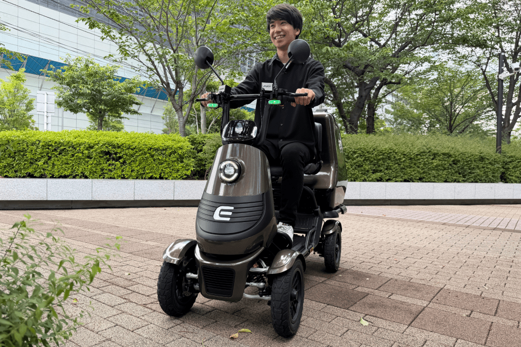 Smiling person rides a brown three-wheeled mobility scooter on a paved path with green trees and a modern building in the background.