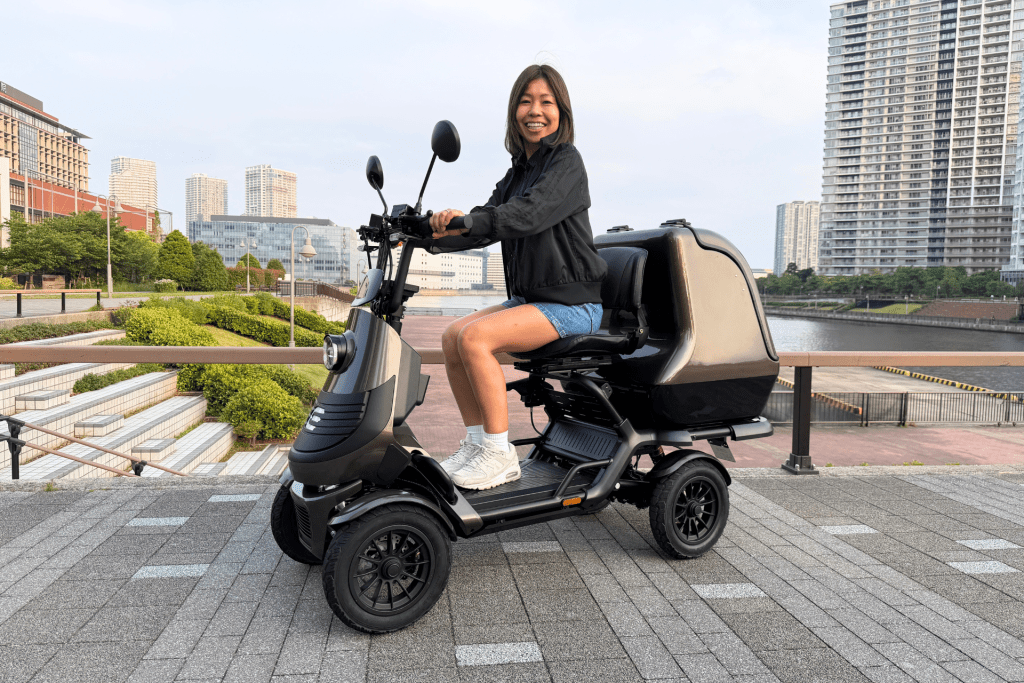 Woman smiling while seated on a black three-wheeled scooter, parked along a riverside promenade with modern buildings in the background.