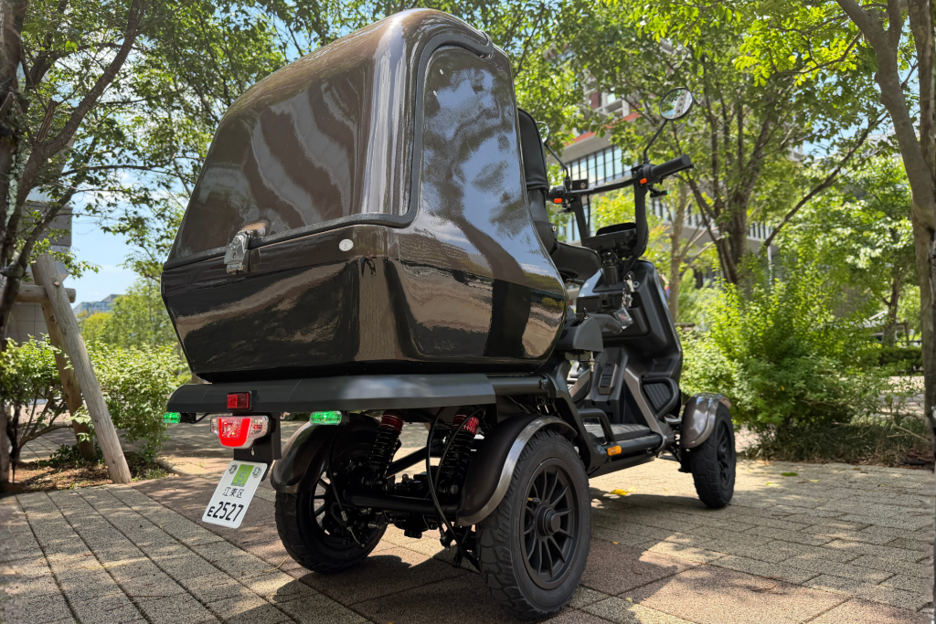 Black motorcycle with a large glossy top case mounted at the rear, parked on a paved path with trees nearby.