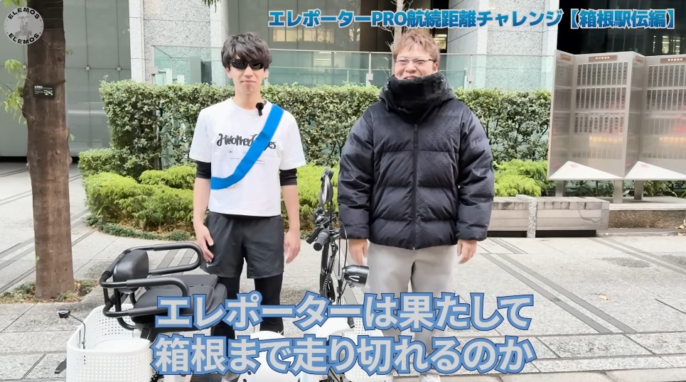 Two men stand outdoors beside a bicycle with a basket, one wearing a white t‑shirt and sunglasses and the other in a black puffy jacket; Japanese video title overlays the scene.