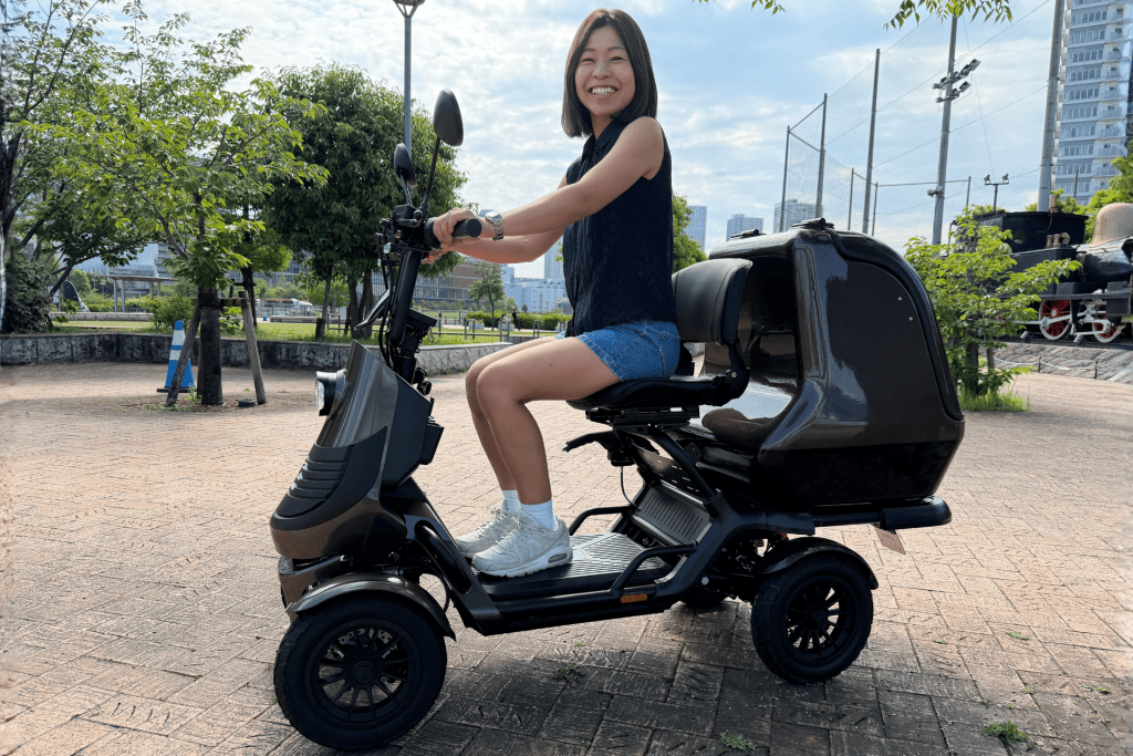 Smiling woman sits on a black scooter in a park, wearing a sleeveless top and denim shorts.