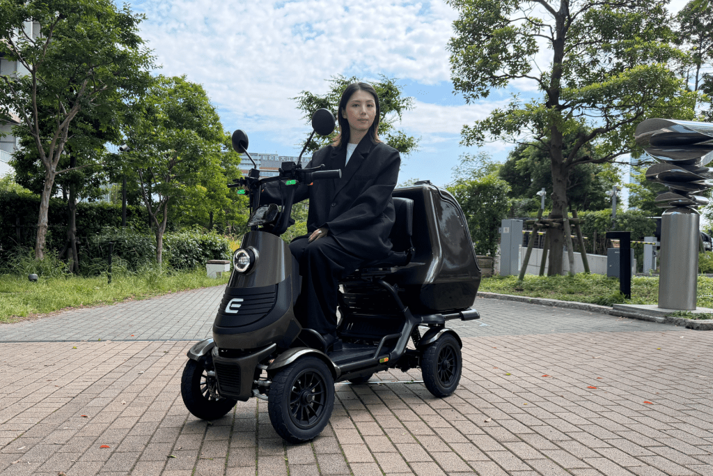 Woman in a black blazer sits on a dark mobility scooter outdoors, with trees and a blue sky in a park setting.