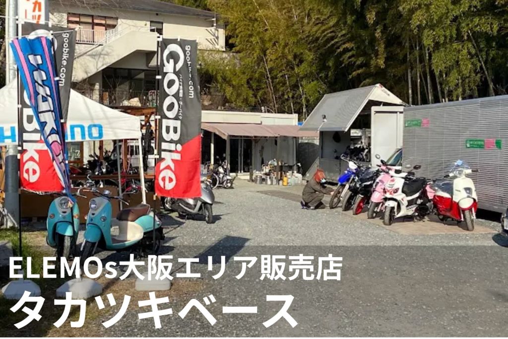 Outdoor scooter shop with colorful vertical banners, multiple motorcycles parked on gravel and a small showroom building in the background, with trees behind.