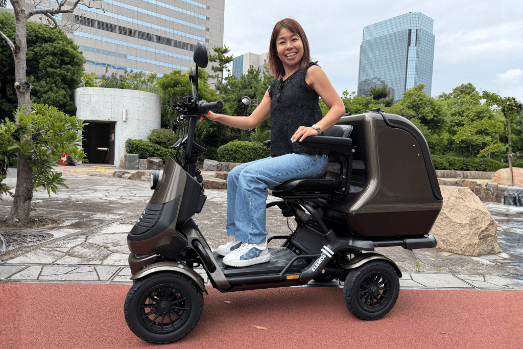 ELEGRAN PREMIUM③ | ELEMOs Smiling woman sits on a brown mobility scooter outdoors in a plaza with greenery and tall buildings in the background, facing the camera.