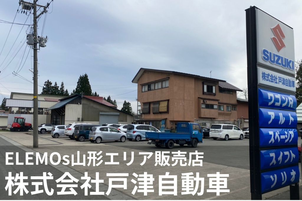 Tall Suzuki dealership sign with blue banners over a parking lot of cars near a beige building under a cloudy sky