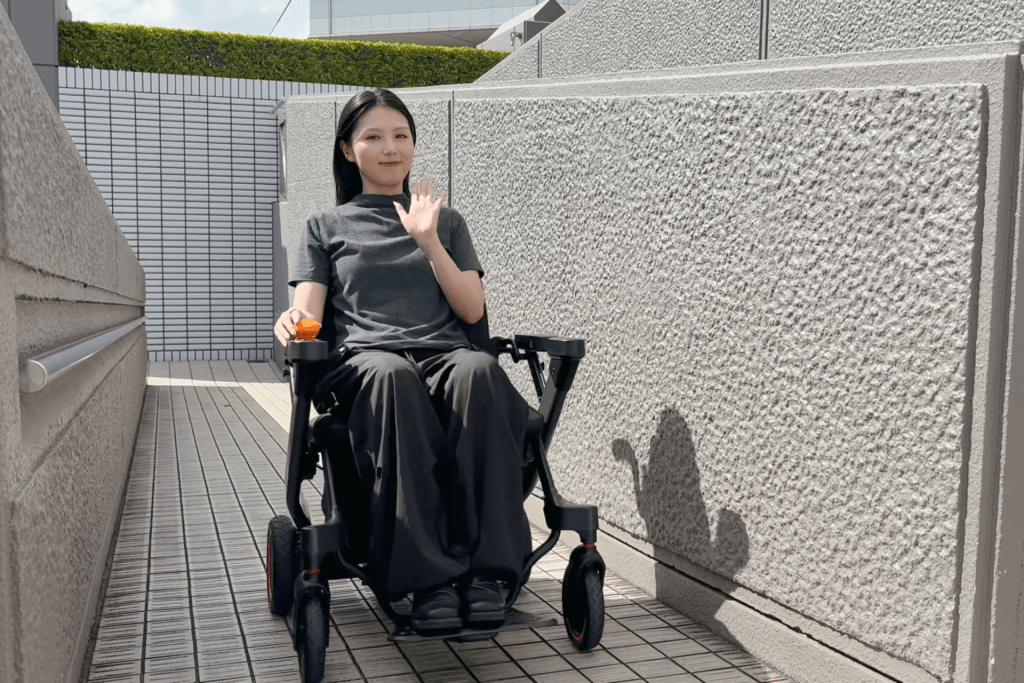 Woman in a wheelchair waving hello down a narrow outdoor walkway with textured concrete walls and tiled floor.