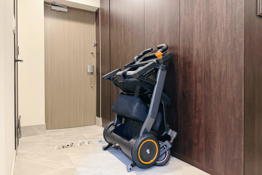 Folded baby stroller with orange accents leaning against a wood-paneled wall in a hallway; shoes on the floor nearby.