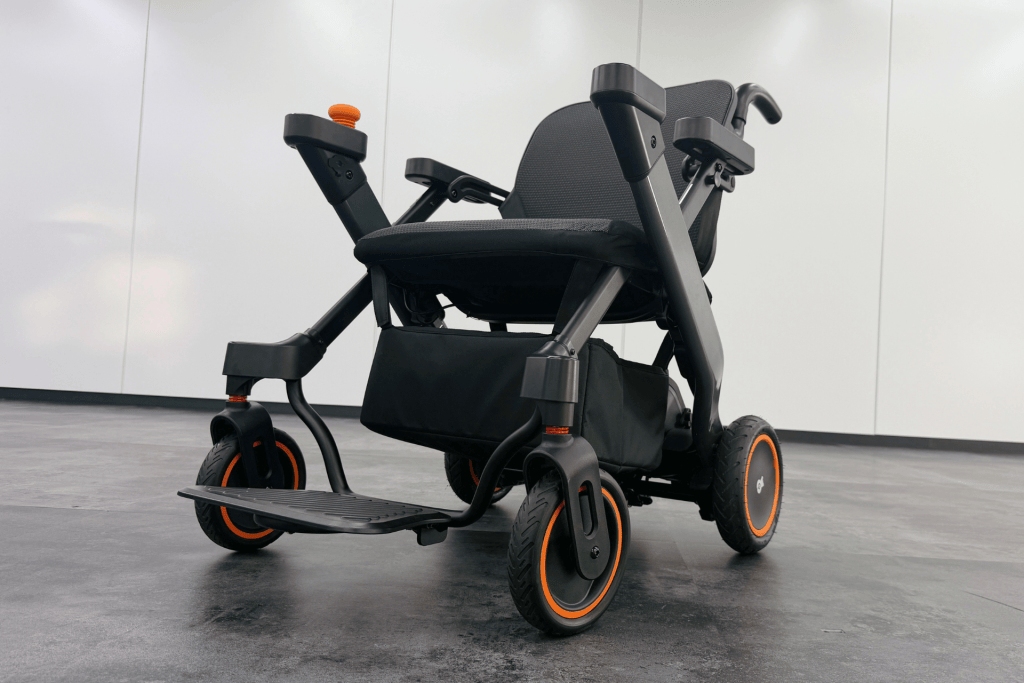 Powered wheelchair with black frame and orange-rimmed wheels, parked indoors on a gray floor.