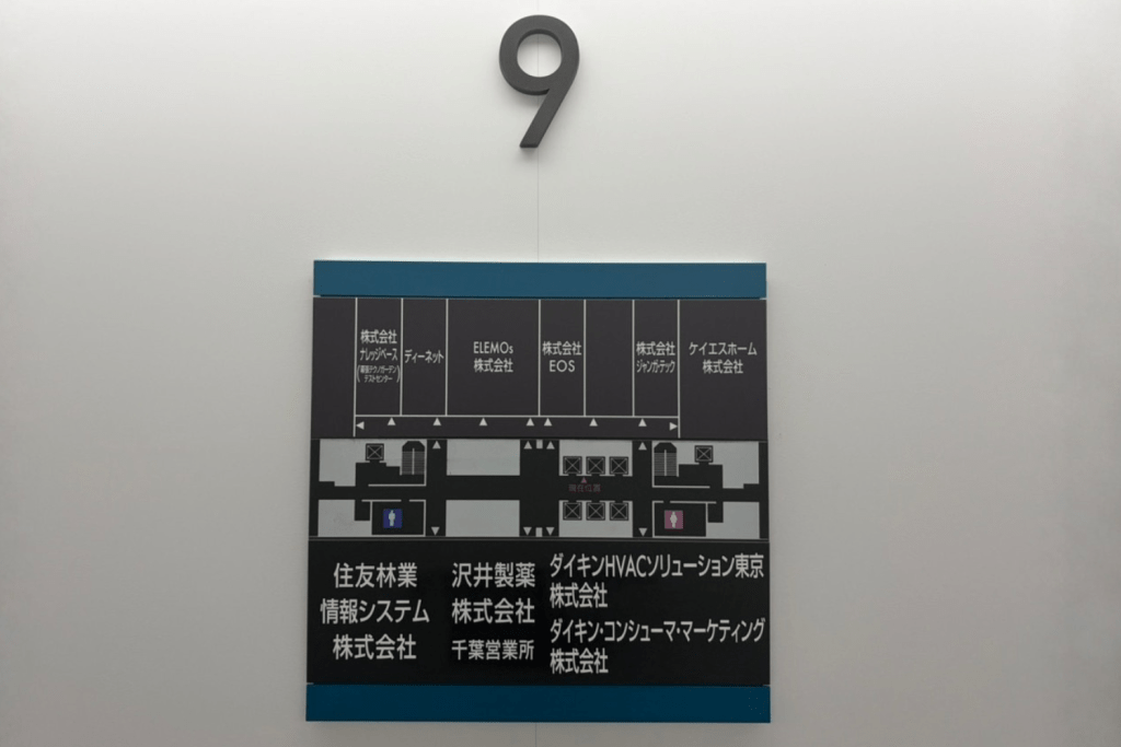 Floor directory sign under a large 9, on a grey wall, listing tenants in Japanese with a simple map layout.