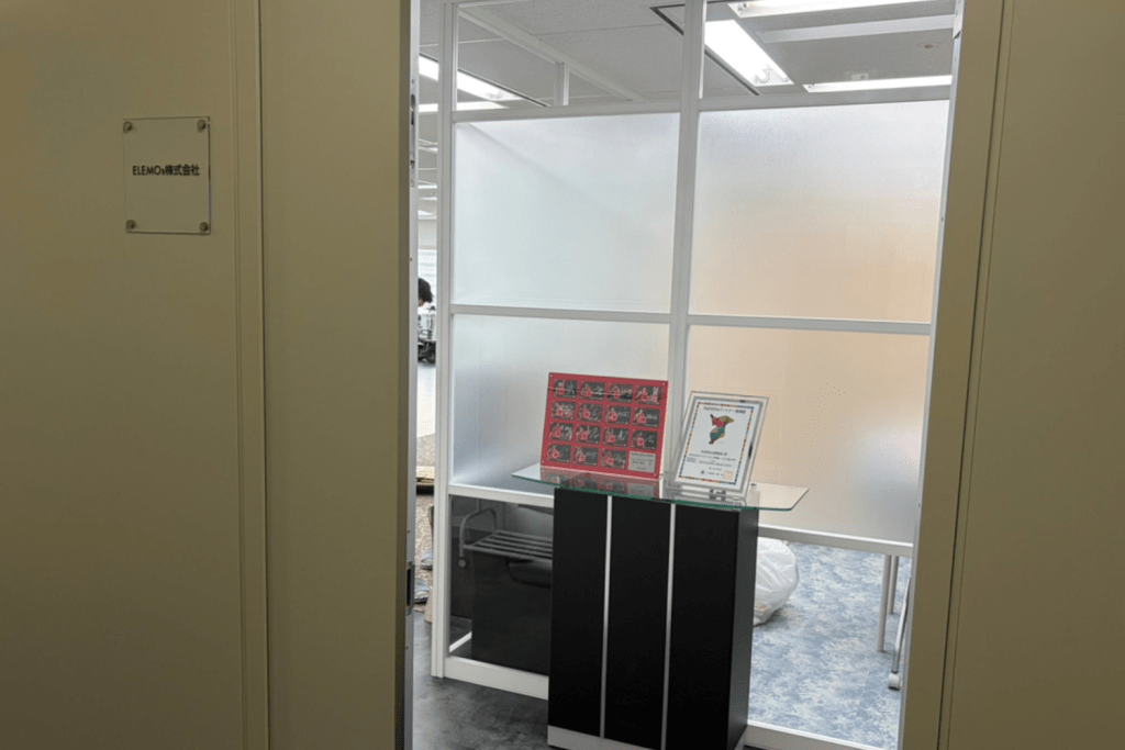 Office entrance to a reception area with a glass display stand and framed posters on a glass-topped pedestal near frosted panels.
