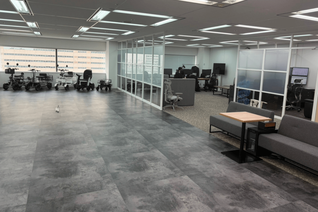 Open office space with several mobility scooters parked by large windows, glass-walled offices in the center, and a gray sofa seating area with a small wooden table on the right side.