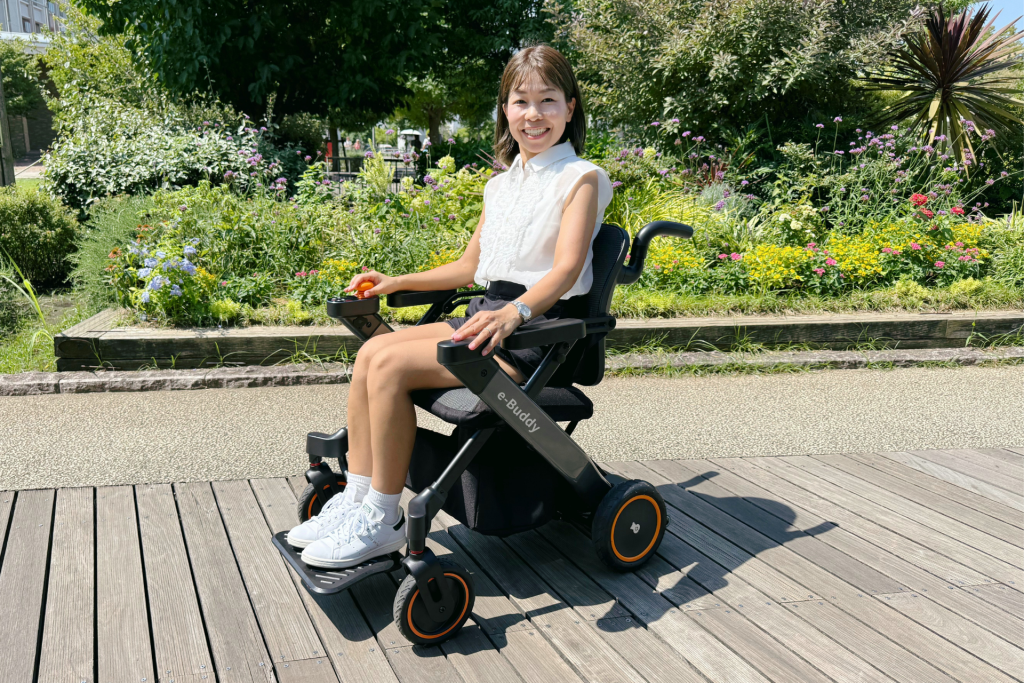 Smiling woman in a white sleeveless blouse sits in a black electric wheelchair on a wooden boardwalk, with a sunny garden full of colorful flowers behind her.