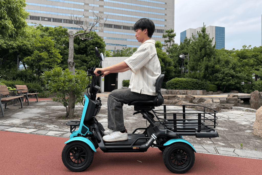 Young man seated on a teal electric scooter with a rear cargo rack, riding in a park with trees and benches nearby.