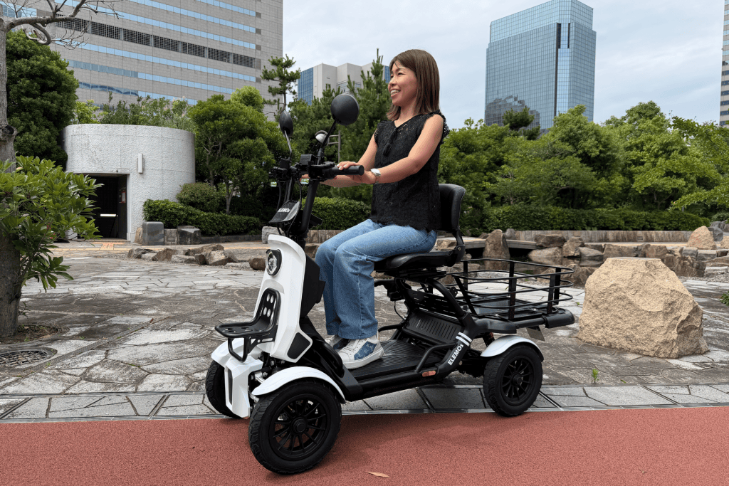 Woman smiling while riding a white three-wheeled mobility scooter in a city park with tall office buildings in the background.