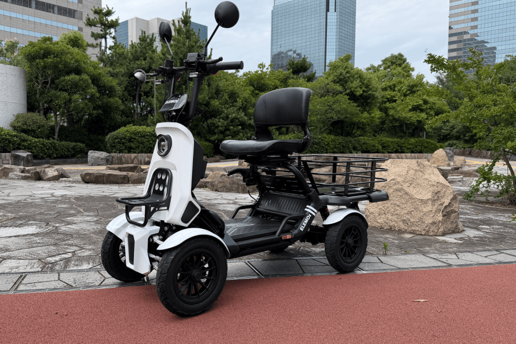 White electric scooter with a black cushioned passenger seat and rear cargo rack parked on a stone plaza, with modern glass buildings in the background.