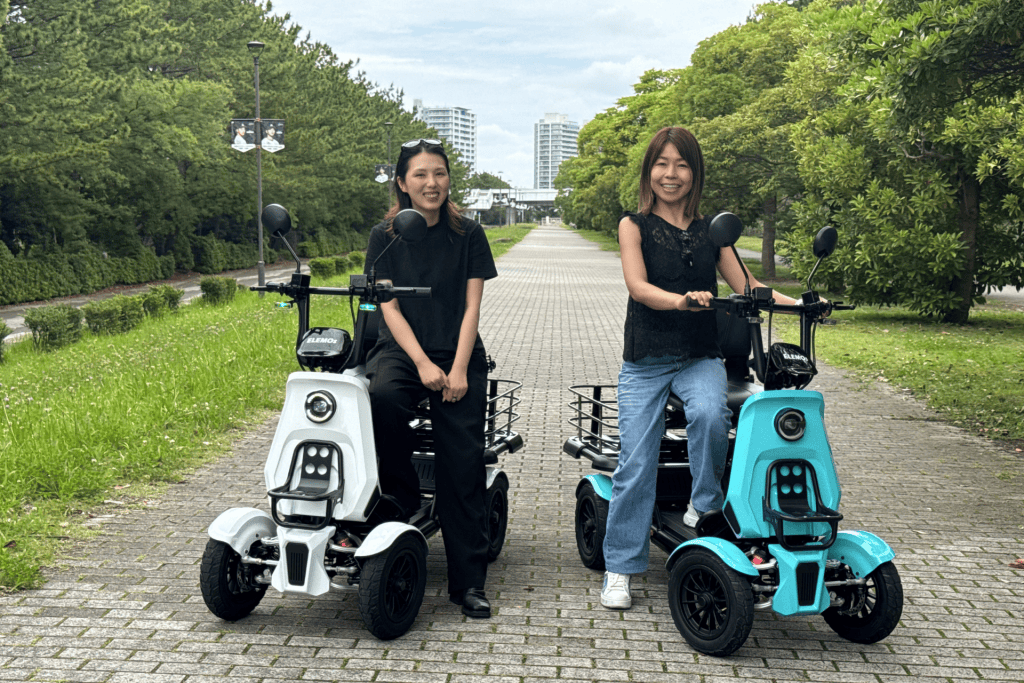 Two women sit on electric scooters on a brick path in a tree-filled park, smiling at the camera.