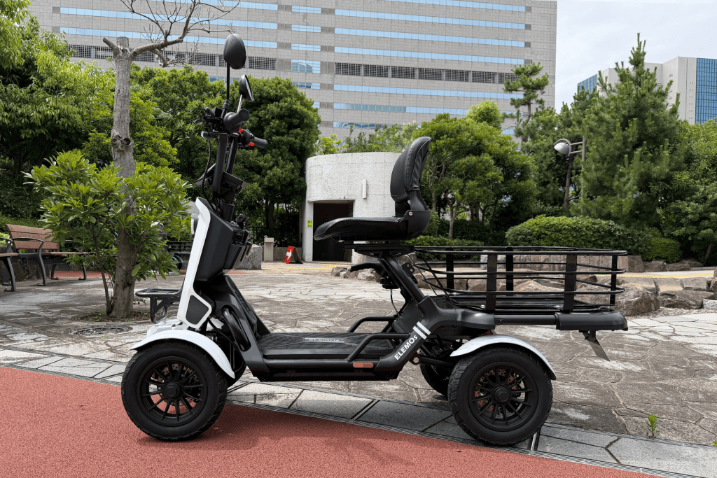 Electric mobility scooter with a large rear cargo rack parked in a park-like area, with trees and a modern building in the background.