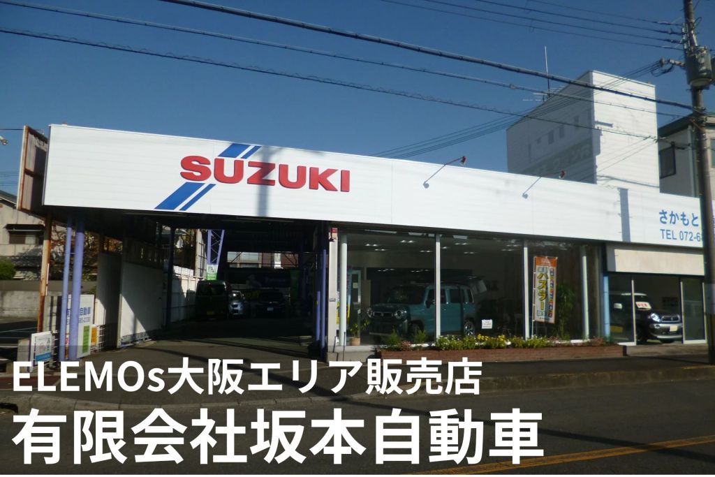 Suzuki dealership storefront with a large white sign and blue diagonal stripes, showing cars inside the showroom under a clear blue sky.